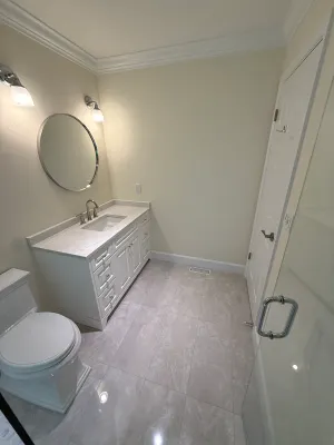 Renovated ensuite with a white vanity, a round mirror, wall sconces, and beige large-format floor tile, by DreamPro Contracting